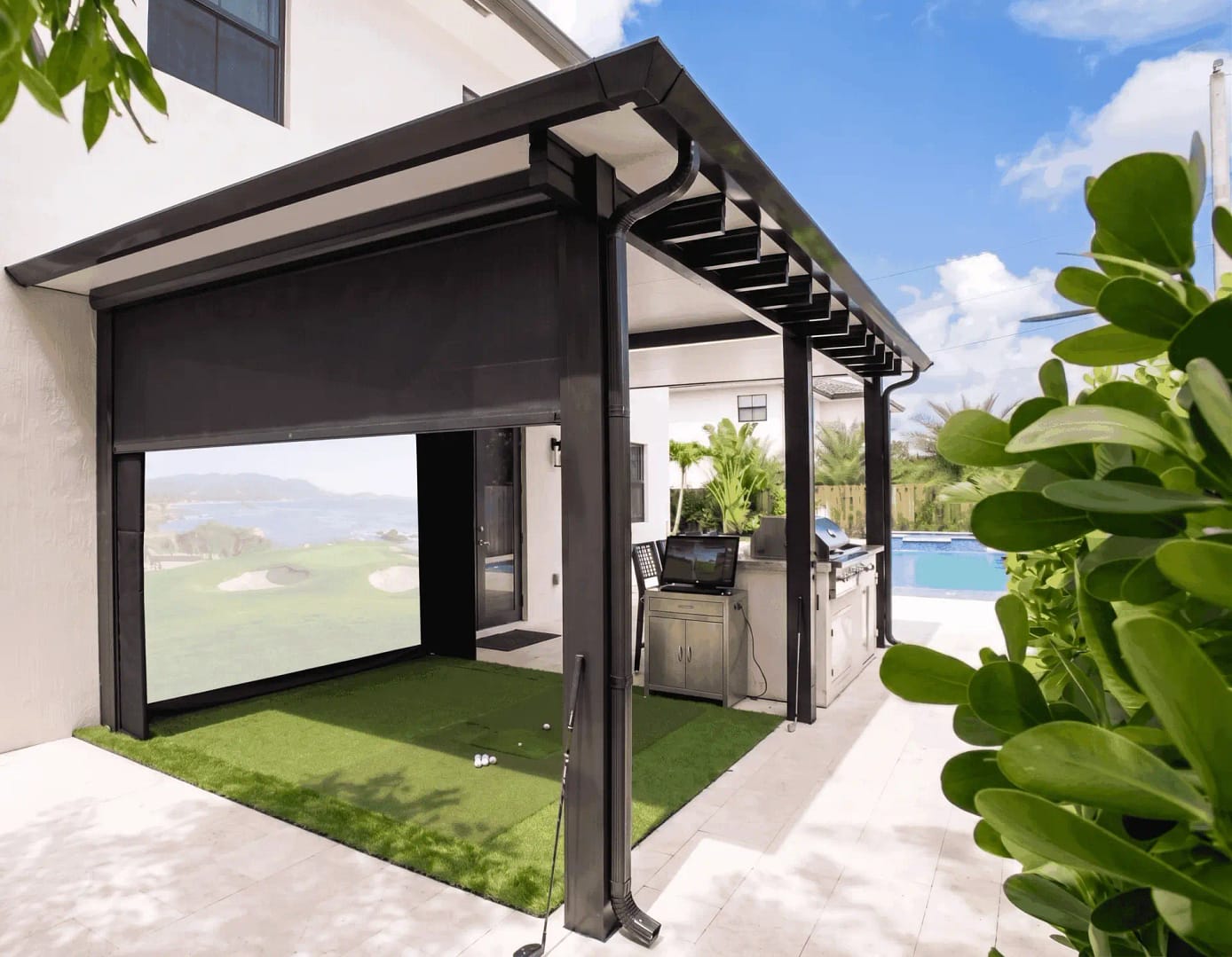 Luxury Outdoor Patio with Custom Golf Simulator and Kitchen - Sims & Greens A residential backyard patio featuring an outdoor golf simulator built into a dark metal pergola structure. The simulator has a deep black enclosure and a large screen displaying a coastal golf course, slightly washed out by the ambient daylight. Next to the green hitting mat is a stainless steel outdoor kitchen, with a blue swimming pool and palm trees in the background under a clear sky.