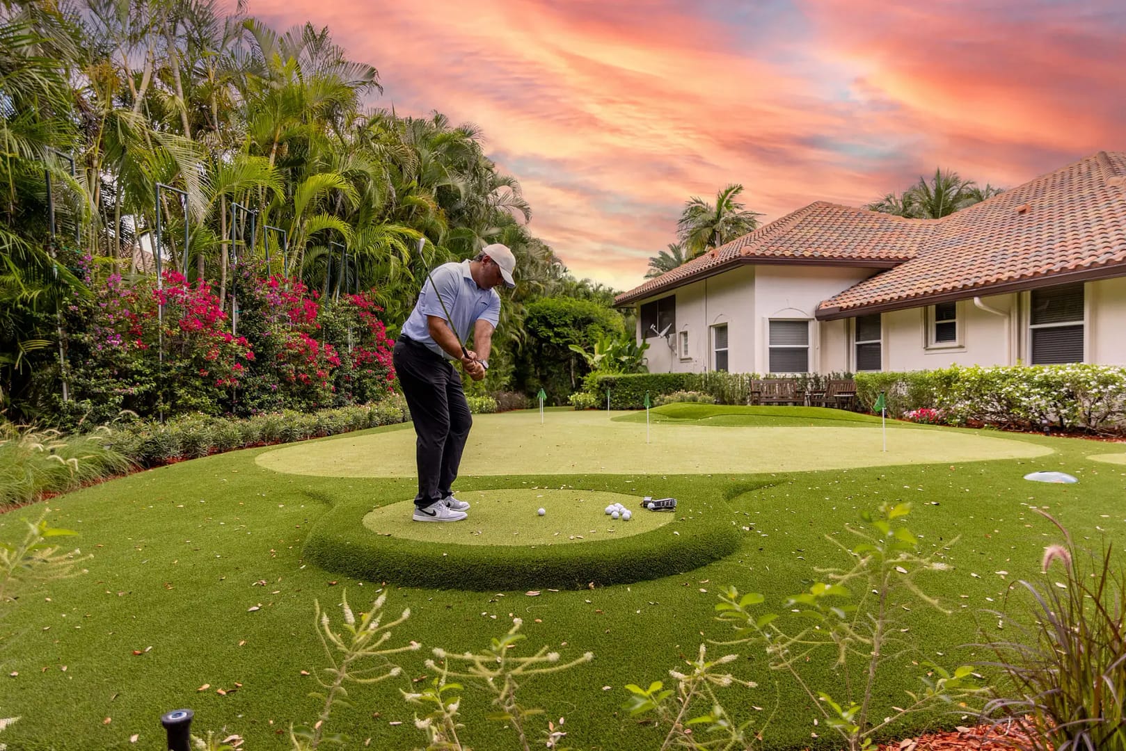 A golfer practicing short game shots on a custom backyard synthetic putting green using a dedicated chipping mat at sunset outside a luxury Belleair, Florida home.