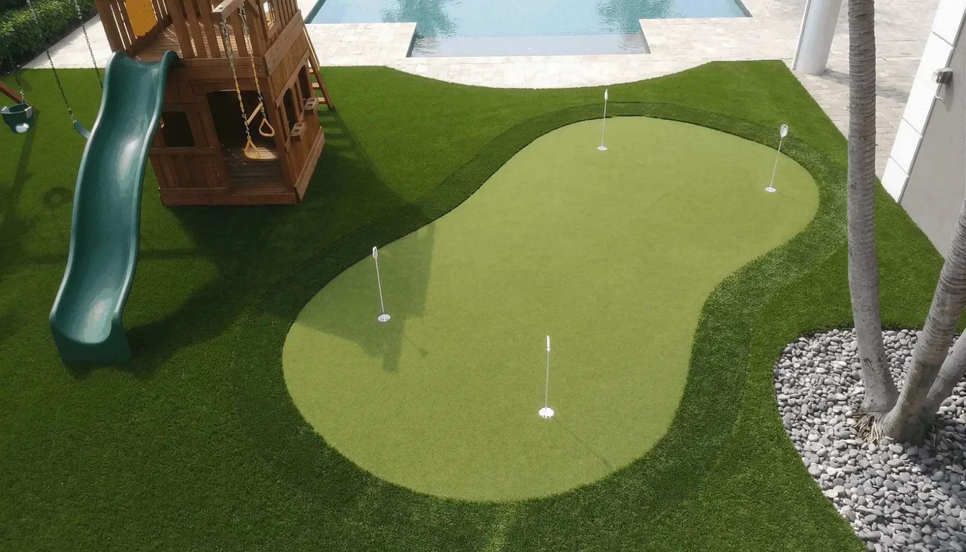 Elevated view of a custom artificial turf putting green with four holes and white flags, located next to a wooden playground structure with a green slide, with a modern swimming pool in the background.