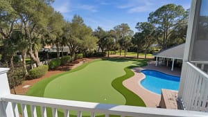 Luxury Florida Backyard Putting Green with Pool | Sims Greens - Sims & Greens Backyard view featuring a curved artificial putting green beside a blue, kidney-shaped pool, with palm trees and a covered patio area in the background.