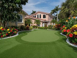 luxury-backyard-putting-green-clearwater-fl - Sims & Greens Luxury backyard artificial turf putting green installation in Clearwater, FL, featuring vibrant tropical landscaping with hibiscus, palm trees, and a custom two-story pink home.