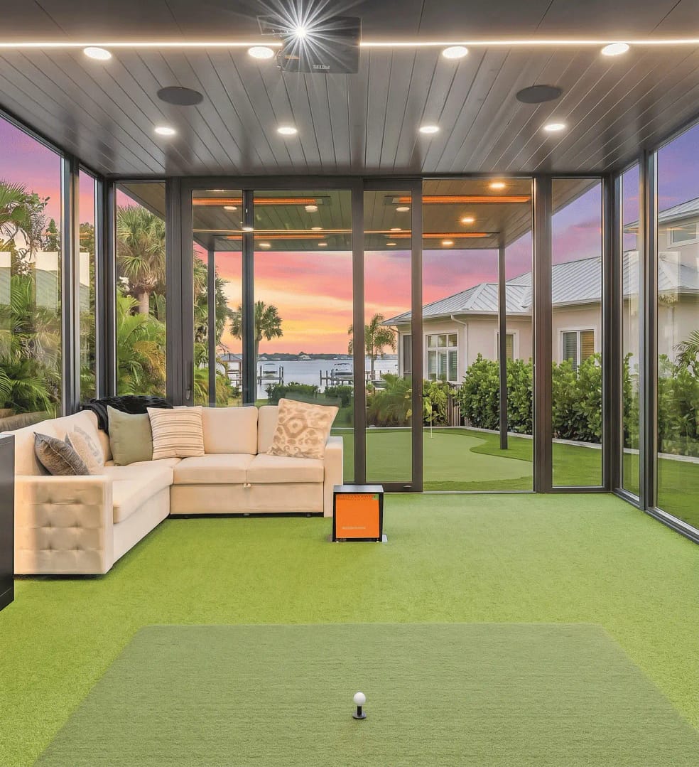 Luxury Indoor Home Golf Simulator with Clearwater Waterfront View - Sims & Greens A spacious indoor golf simulator room with a white L-shaped couch on the left and a golf hitting mat in the foreground. Large glass doors open to a backyard putting green and a view of a tranquil Florida waterway at sunset.